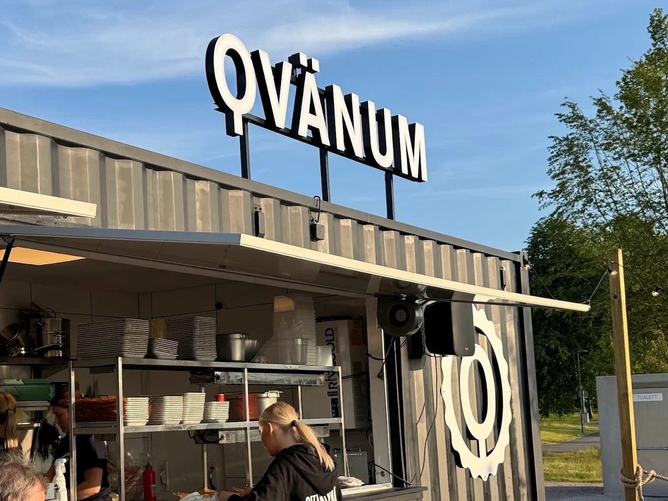 En serveringscontainer med skylten Qvänum på taket under en klarblå himmel.