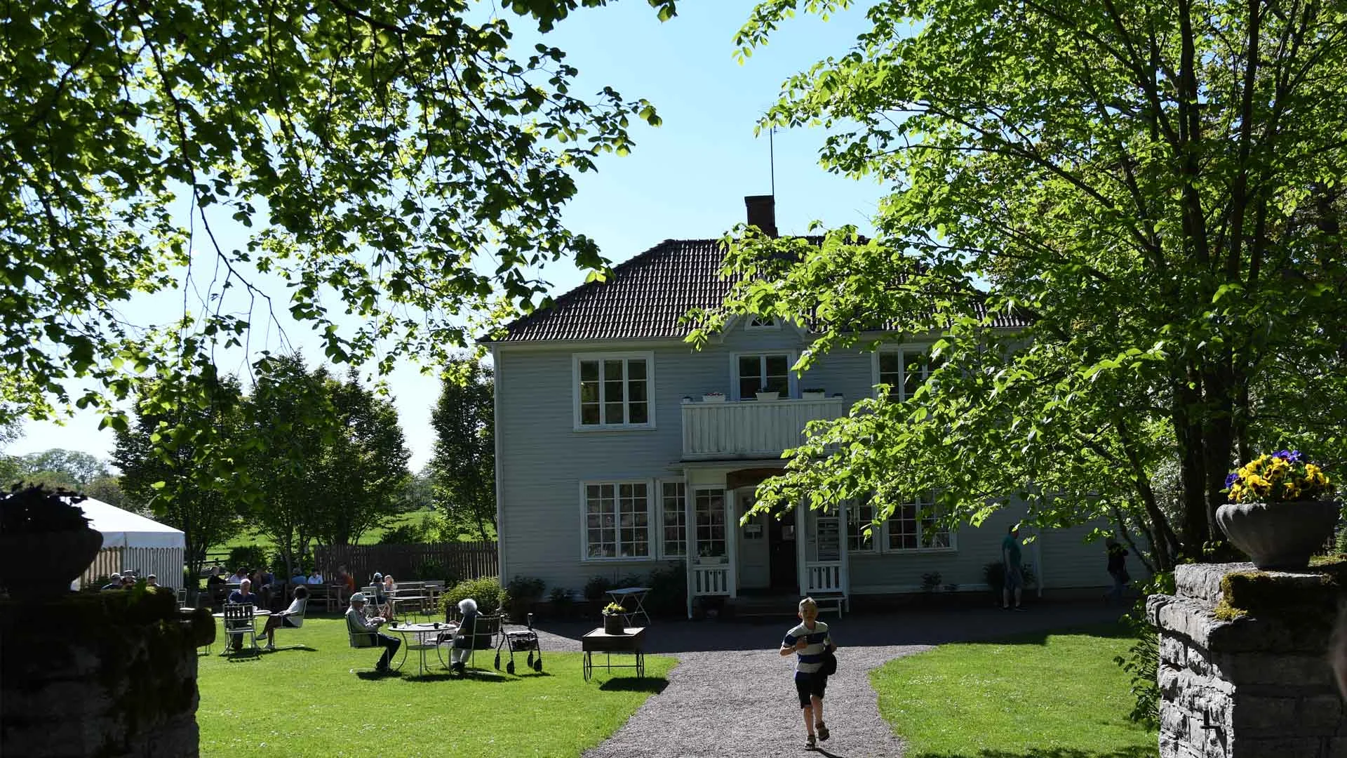 Sommargäster som fikar utomhus vid Varnhems Klostercafé med utsikt mot klosterkyrkan i Skara kommun.